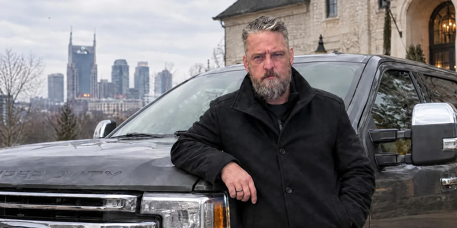 Shannon Higgason, grey-bearded and in a black coat, leaning on the front of a pickup truck with a city skyline behind him.